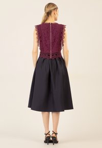 Burgunderfarbenes Spitzenoberteil mit gewellten Kanten, kombiniert mit einem schwarzen, fließenden A-Linien-Rock. Das Modell trägt schwarze, römische Absätze. Rückansicht.