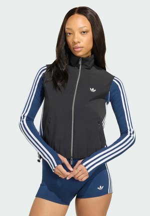 Młoda kobieta ubrana w czarną kamizelkę na zamek, na niebieskim dresie Adidas z białymi paskami, stojąca z złączonymi rękami.