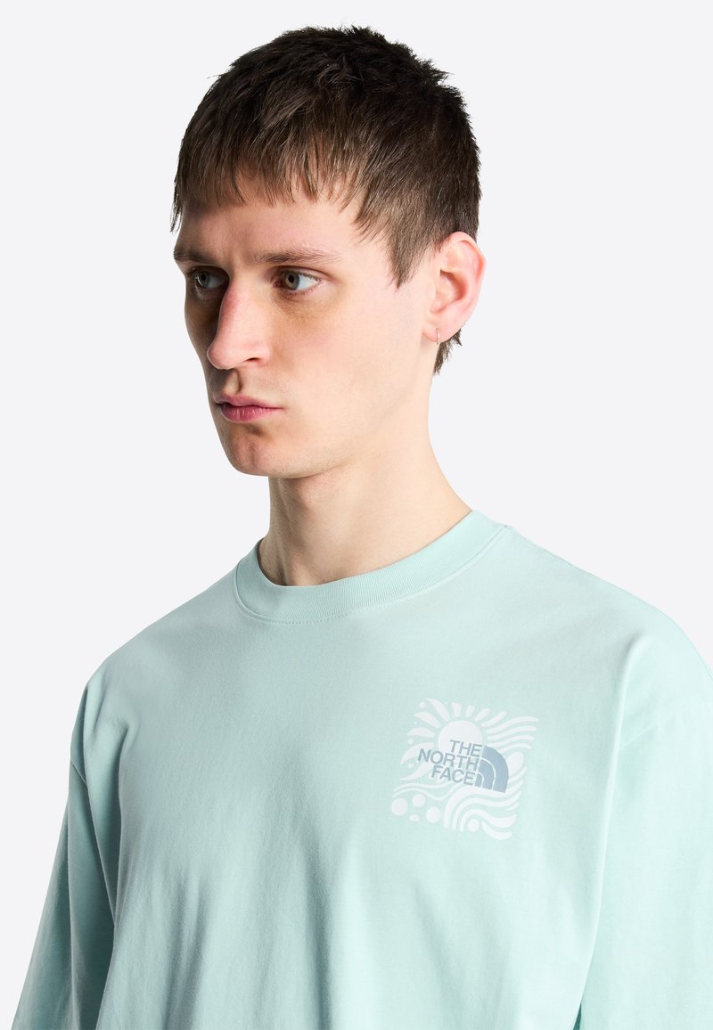 Junger Mann mit kurzen braunen Haaren, der einen hellblauen The North Face Pullover trägt und auf einfachem Hintergrund nach links schaut.