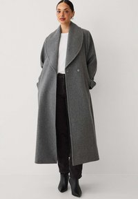 Lang grå uldblandet frakke med sjalskrave, dobbeltknaplukning og sidelommer, båret over en hvid top og sorte jeans.
