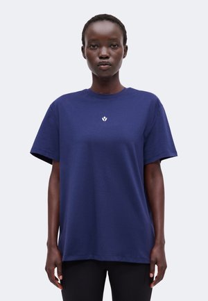Femme aux cheveux courts portant un t-shirt bleu marine uni avec un petit logo blanc au centre, debout devant un fond clair.