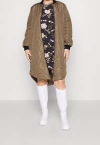 Persona que lleva un abrigo acolchado de oliva hasta la rodilla, abierto sobre un vestido floral negro, combinado con botas blancas hasta la rodilla, contra un fondo liso.