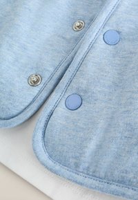 Tessuto leggero blu chiaro, morbido al tatto, con bottoni a pressione argentati e un pulsante blu di accento. Presenta bordi arrotondati e un tessuto bianco contrastante al di sotto.