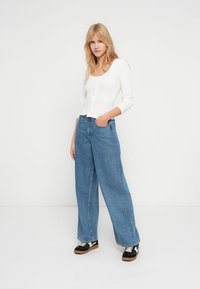 Λευκή μπλούζα με μακριά μανίκια και κουμπιά, σε συνδυασμό με φαρδιά μπλε τζιν. Το μοντέλο φοράει μαύρα αθλητικά παπούτσια με λευκές λεπτομέρειες. Απλός, καθημερινός σχεδιασμός.