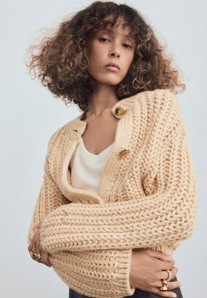 Persona joven con cabello rizado que lleva puesto un suéter de punto grueso color beige, una blusa blanca y anillos dorados en los dedos, contra un fondo sencillo.