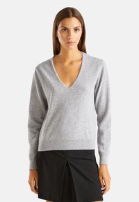 Šedý svetr s výstřihem do V vyrobený z měkké tkaniny, s dlouhými rukávy a žebrovaným lemem. Vytvořený v kombinaci s černou sukní. Minimalistický design.