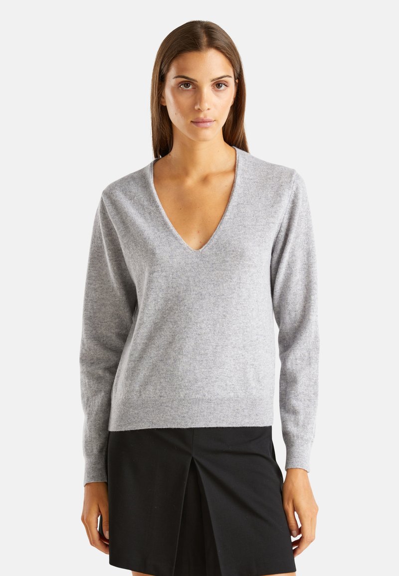 Šedý svetr s výstřihem do V vyrobený z měkké tkaniny, s dlouhými rukávy a žebrovaným lemem. Vytvořený v kombinaci s černou sukní. Minimalistický design.