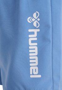 Zbliżenie na jasnoniebieską tkaninę z pionowo nadrukowaną białą nazwą marki „hummel” i logo pszczoły.