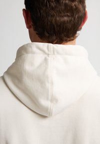 Ecrufarbenes Kapuzensweatshirt mit strukturierter Oberfläche, das ein einfaches Design und sichtbare Nähte entlang des Kragens und der Kapuze aufweist.