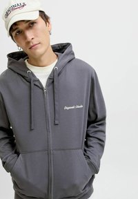 Sudadera gris con cremallera, cordones, bolsillos frontales y texto bordado "Originals Studio". Tejido suave y corte casual.
