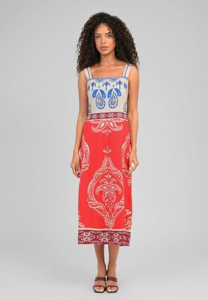 Femme aux cheveux bouclés portant une robe sans manches avec un haut à motif bleu et une jupe rouge avec des motifs floraux crème, debout face à l'avant.