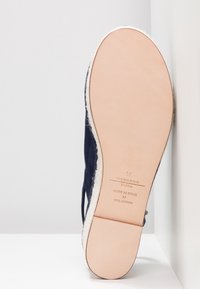Chaussure en daim bleu marine foncé avec doublure blanche en fourrure, semelle en cuir beige et texte imprimé au fond. La forme est arrondie avec un design décontracté.