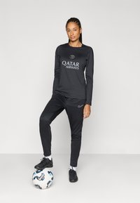 Fekete hosszú ujjú sportfelső "QATAR AIRWAYS" logóval, fekete nadrággal párosítva. Fekete focicipő, mintázott focilabdán állva.