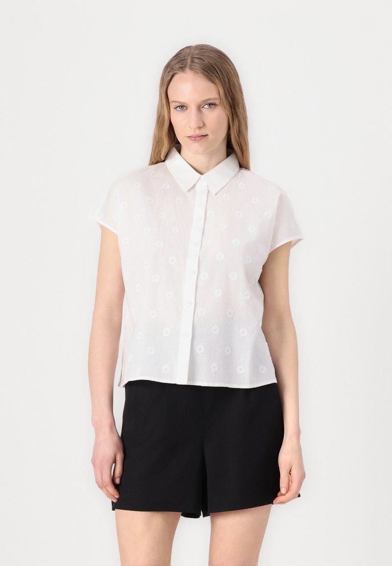 Camicia bianca a maniche corte con chiusura a bottoni, caratterizzata da ricami floreali e colletto. Abbinata a pantaloni corti neri.