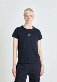 Navy blå bomull T-skjorte med rund hals og korte ermer; har en logo brodert i hvitt på brystet. Myk tekstur, avslappet passform.