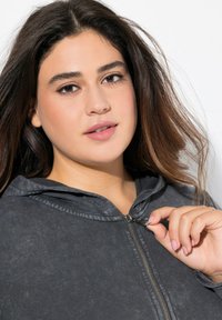 Dunkelgrauer Reißverschluss-Hoodie aus strukturiertem Material, mit Kapuze und Metallreißverschluss. Das Model hält den Reißverschluss mit einer Hand.