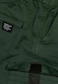 Tessuto verde scuro con una texture liscia; presenta un'etichetta nera con "SURPLUS" e un dettaglio di chiusura in Velcro sul pannello frontale.