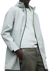 Chaqueta impermeable gris claro con capucha, ribetes negros, dos bolsillos laterales y un ajuste relajado, llevada sobre una camisa blanca y pantalones caqui.
