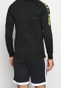 Sudadera atlética negra con un respaldo texturizado, presenta acentos verdes con patrones en las mangas, combinada con pantalones cortos negros con ribetes blancos.