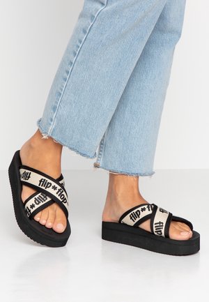 Piedi che indossano sandali platform neri con cinturini beige incrociati etichettati "flip flop,", abbinati a jeans azzurri chiaro con orlo sfrangiato su sfondo bianco.