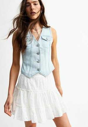 Chaleco de denim azul claro con un diseño ajustado, cinco botones y dos bolsillos en el pecho, combinado con un vestido blanco de capas con bordado.