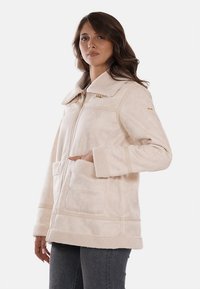 Giacca in pile beige con colletto, zip frontale e due tasche. Presenta una texture morbida e dettagli di cucitura lungo le cuciture.
