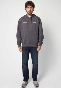 Sweat à capuche gris avec poche frontale et corde de serrage, présentant un texte bleu et blanc. Associé à un jean bleu foncé et des baskets grises.