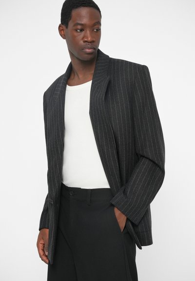 Ciemnoszary żakiet w prążki z poduszanymi ramionami, jedynym zapięciem na guzik, szerokimi klapami oraz luźnym krojem, noszony na białej koszuli.