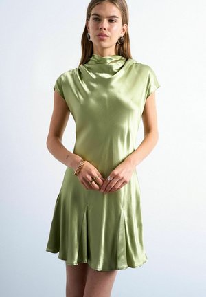 Kvinde iført ærmeløs lysegrøn satin kjole med plisseret nederdel, accessoriseret med guld armbånd, ringe og runde øreringe, stående mod en hvid væg.