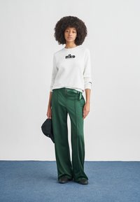 Γκρι φούτερ με μαύρο λογότυπο Nike, συνδυασμένο με πράσινο σατέν παντελόνι με φαρδιά μπατζάκια. Το μοντέλο κρατάει ένα μαύρο καπέλο και στέκεται μπροστά σε ένα λευκό φόντο.