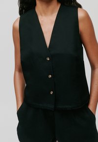Czarna bezrękawnik z dekoltem w kształcie litery V, ozdobiony pięcioma metalowymi guzikami i skróconym krojem. Materiał ma gładką teksturę i postrzępione brzegi.