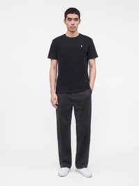 Stående mann iført en svart t-skjorte med korte ermer, mørkegrå bukser og hvite sneakers mot en ensfarget hvit bakgrunn.