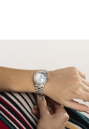 Personne ajustant une montre-bracelet en métal argenté avec cadran blanc et affichage de la date, portant un vêtement textile rayé multicolore.