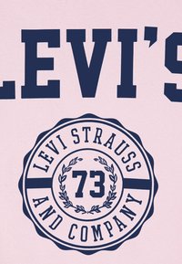 Le texte bleu marine indique "LEVI'S" au-dessus d'un logo circulaire avec "LEVI STRAUSS AND COMPANY" et "73," sur un fond rose.