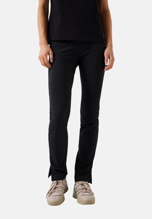 Persoon draagt zwarte slim-fit broek en lichtbeige sneakers, staand tegen een effen lichte achtergrond.