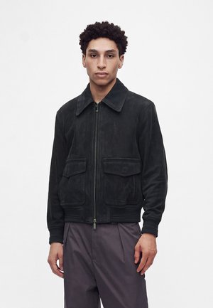 Junger Mann mit kurzen lockigen Haaren, trägt eine schwarze Jacke mit Reißverschluss und zwei vorderen Klappentaschen sowie dunkelgraue Hosen, blickt nach vorne.