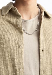 Beige texturiertes Hemd mit Knöpfen und Kragen, getragen über einem hellgrauen T-Shirt, ergänzt durch eine silberne Kettenhalskette.