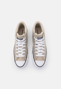 Béžové plátěné tenisky s bílými tkaničkami, gumovou špičkou a černými akcenty. Obsahují vnitřní branding a strukturovaný povrch.