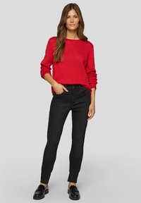 Rot strukturierter Pullover mit weitem Ausschnitt, kombiniert mit engen schwarzen Jeans und schwarzen Slippern. Das Model steht vor einem neutralen Hintergrund.