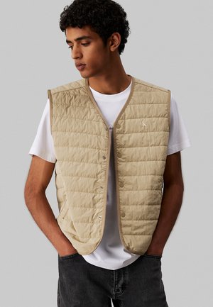 Beige gepolsterte Weste mit gestepptem Design, Druckknopfverschluss und seitlichen Taschen, getragen über einem weißen Hemd. Verfügt über dezentes Branding in der oberen linken Ecke.