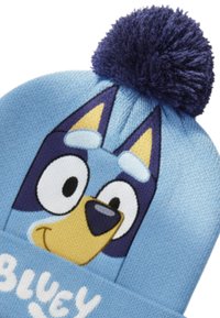 Beanie lavorato a maglia blu con un design di faccia di cane cartoon in tonalità di blu e giallo, con un pom pom blu scuro sulla parte superiore e il testo "BLUEY".