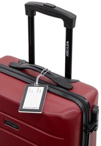 Red hardshell suitcase with a textured surface, black zippers, and a retractable handle. Features a luggage tag attached to the zipper.