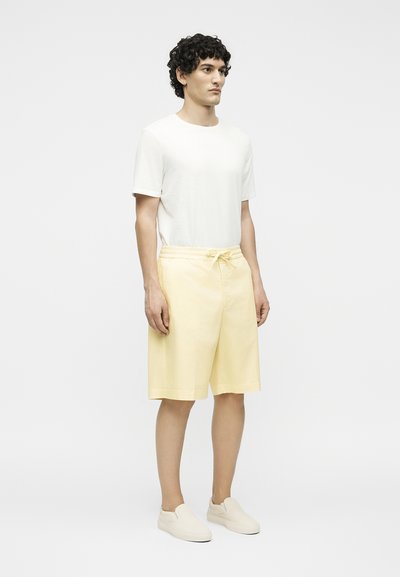 Άνδρας με σγουρά μαλλιά στέκεται φορώντας λευκό κοντομάνικο μπλουζάκι, ανοιχτά κίτρινα σορτς μέχρι το γόνατο με κορδόνι και κρεμ παπούτσια χωρίς κορδόνια.