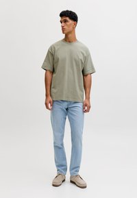 Bărbat în picioare, purtând un tricou larg verde salvie, blugi albaștri deschiși și încălțăminte mocasin bej, pe un fundal simplu și deschis la culoare.