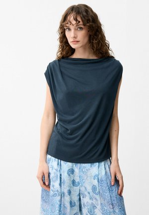 Kvinde med krøllet hår iført en mørk navy ærmeløs top og lyseblå vidde bukser med paisley-mønster mod en hvid baggrund.