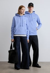 Hellblaue Kapuzenpullover mit Kordelzügen und Fronttaschen, kombiniert mit dunklen, weitem Jeans. Eine schwarze Stofftasche mit einem Logodetail.