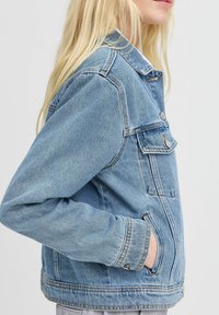 Chaqueta de mezclilla azul con cuello, bolsillos laterales y detalles de botones. Presenta un diseño clásico con un ajuste ligeramente holgado y costuras visibles.