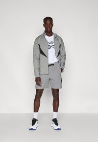 Lichtgrijs sportjack met zwarte accenten, gecombineerd met bijpassende shorts. Witte T-shirt eronder met een logo. Zwarte en witte sneakers.