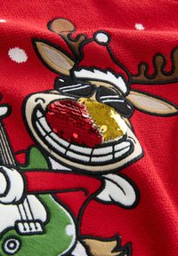 Rødt strikket stof med et cartoon-rendyr iført en julestråhat. Rensdyret har pyntet sin næse med røde og gyldne palietter og holder en grøn guitar.