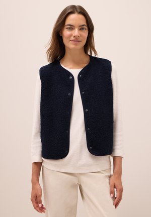 Gilet en polaire bleu marine avec un col rond et une fermeture à boutons-pression. Présente un tissu texturé et une coupe ajustée, porté par-dessus un haut léger.